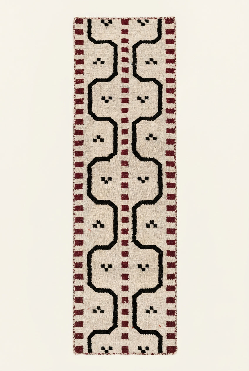 A long rectangular hand woven runner rug with a geometric pattern, The rug has a cream base with black zigzag lines, red dotted borders, and small black motifs.