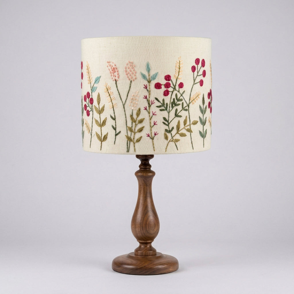 A table lamp with a cream-colored fabric drum shade decorated with hand embroidered pink, red, and green floral motifs.