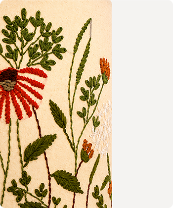 Close-up of a textile or rug featuring embroidered wildflowers on a cream background, with green leaves, a red flower, orange buds, and a white flower cluster.