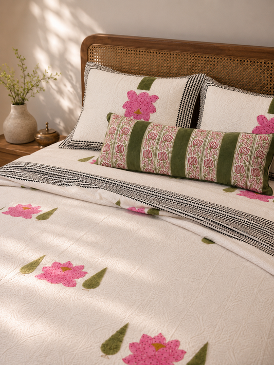 A styled bed with a cream-colored jacquard bedcover and matching pillowcases featuring pink floral motifs and green leaf accents, styled with a striped and hand block printed  decorative sham lumbar pillow. 