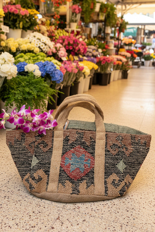 A large woven tote bag with a geometric pattern, featuring natural jute handles and a central red, blue, and beige diamond motif.
