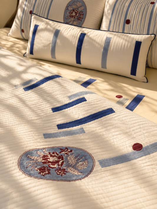 A quilted cream bedding set on a bed, featuring blue and gray striped pillows and an embroidered floral oval motif in blue, red, and beige.