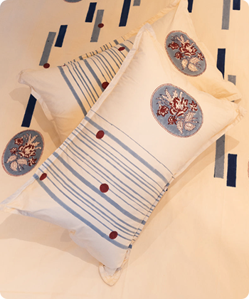A decorative rectangular throw pillow with blue stripes and embroidered floral medallions, resting on a matching patterned cream bedding set.