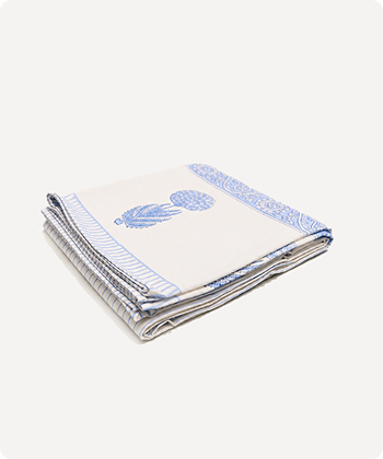 A folded white cotton table linen or napkin with a blue floral print and a blue patterned border, shown on a light off-white background.