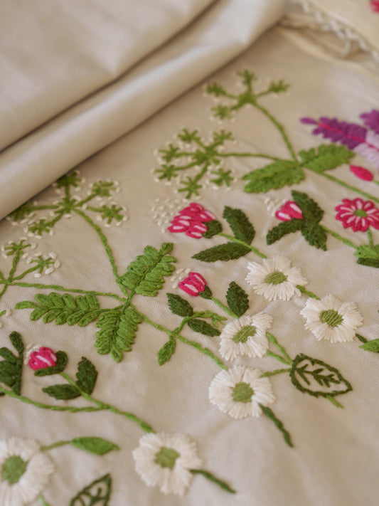 Close-up of a cream-colored embroidered textile with floral patterns in green, white, pink, and purple, with a folded matching fabric roll at the top.
