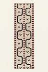 A long rectangular hand woven runner rug with a geometric pattern, The rug has a cream base with black zigzag lines, red dotted borders, and small black motifs.