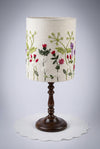 A table lamp shade hand embroidered on a cream fabric drum shade decorated with embroidered green leaves and red, pink, and purple flowers.