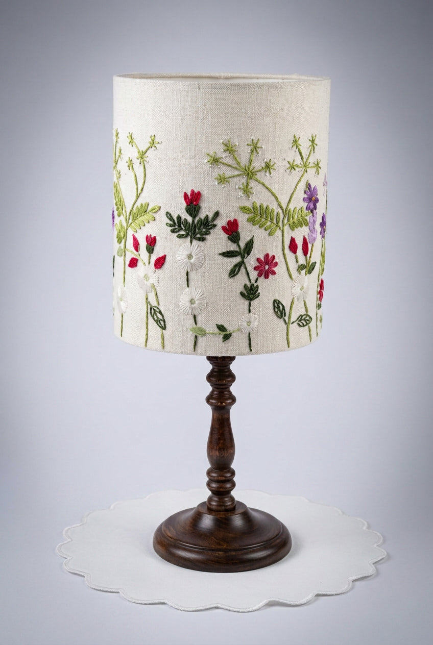 A table lamp shade hand embroidered on a cream fabric drum shade decorated with embroidered green leaves and red, pink, and purple flowers.