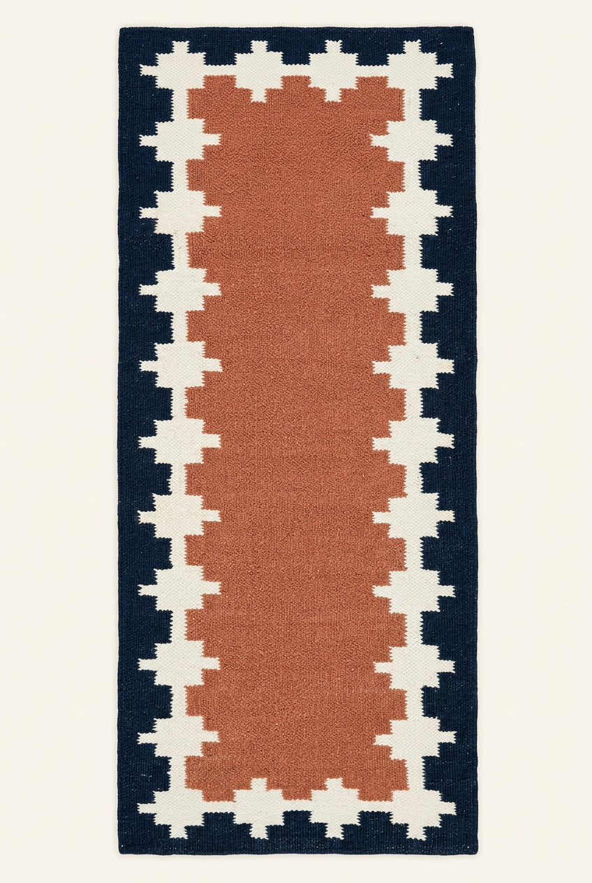 A long rectangular hand woven woolen  runner rug laid flat. The rug has a terracotta pink center, cream geometric borders, and a dark navy outer border.