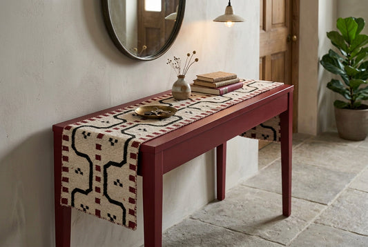 A long rectangular hand woven woolen kilim table runner rug with a geometric pattern, laid flat on a entryway table. The rug has a cream base with black zigzag lines, red dotted borders, and small black motifs.