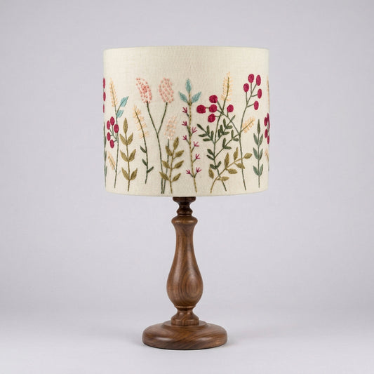 A table lamp with a cream-colored fabric drum shade decorated with hand embroidered pink, red, and green floral motifs.
