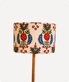 A drum lampshade with a colorful folk-art style pattern featuring red pomegranates, blue floral elements, and green leaves on a cream background, supported by a wooden stand against a light gray background.