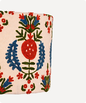 A close-up of a cylindrical container or vase with a cream-colored fabric cover featuring a colorful embroidered or printed floral pattern with red, blue, and green motifs.