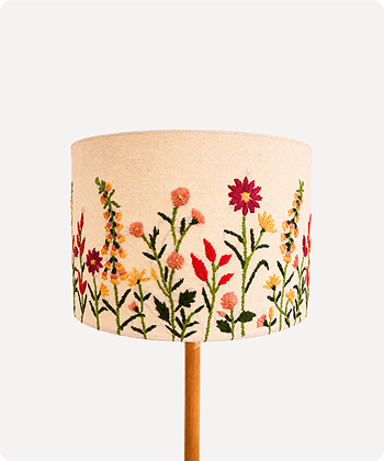 A drum lampshade with a beige linen-like base and colorful embroidered wildflowers, shown on a light wood stand against a plain light gray background.