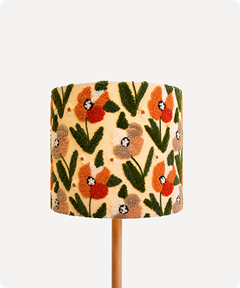 A cylindrical table lamp shade with a cream background and a dense floral pattern featuring orange-red flowers, green leaves, and beige seed heads, mounted on a light wood stem against a light gray background.