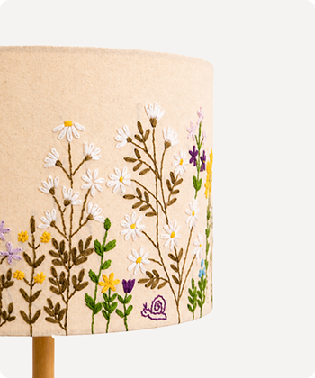 Close-up of a beige fabric lampshade with embroidered wildflowers and green leaves, mounted on a light wood stem against a plain light background.