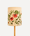 A cylindrical table lamp shade with a natural beige linen-like fabric, decorated with embroidered red flowers and green foliage, mounted on a light wood stand against a plain white background.