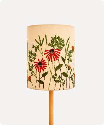 A cylindrical table lamp shade with a natural beige linen-like fabric, decorated with embroidered red flowers and green foliage, mounted on a light wood stand against a plain white background.